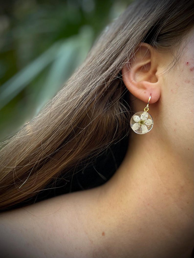 Puede incluir: Un par de pendientes de aro dorados con una flor blanca prensada dentro de un c&iacute;rculo de resina transparente.
