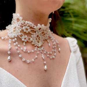 May include: A white beaded choker necklace with a delicate floral design and dangling pearl accents.
