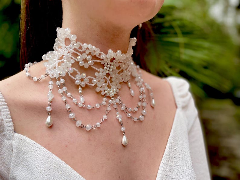 May include: A white beaded choker necklace with a delicate floral design and dangling pearl accents.