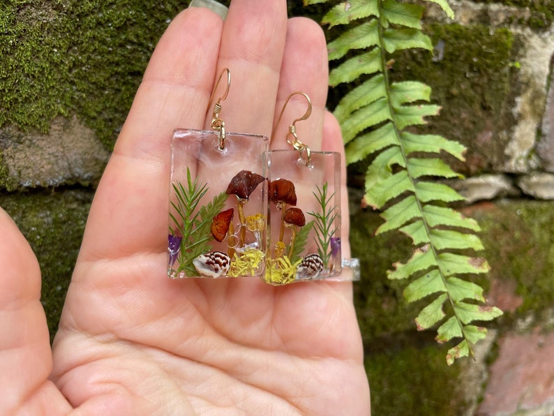 May include: A pair of gold dangle earrings with clear resin squares containing pressed flowers, mushrooms, and seashells.