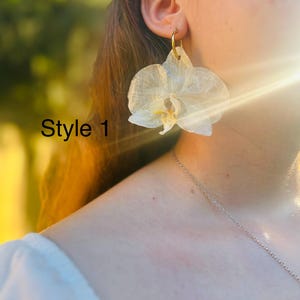 Pode incluir: Um brinco de flor de orqu&iacute;dea branca transl&uacute;cida com um gancho de argola de metal dourado. A estrutura das p&eacute;talas &eacute; vis&iacute;vel com nervuras delicadas e um centro amarelo. O texto Style 1 est&aacute; impresso em preto no lado esquerdo.