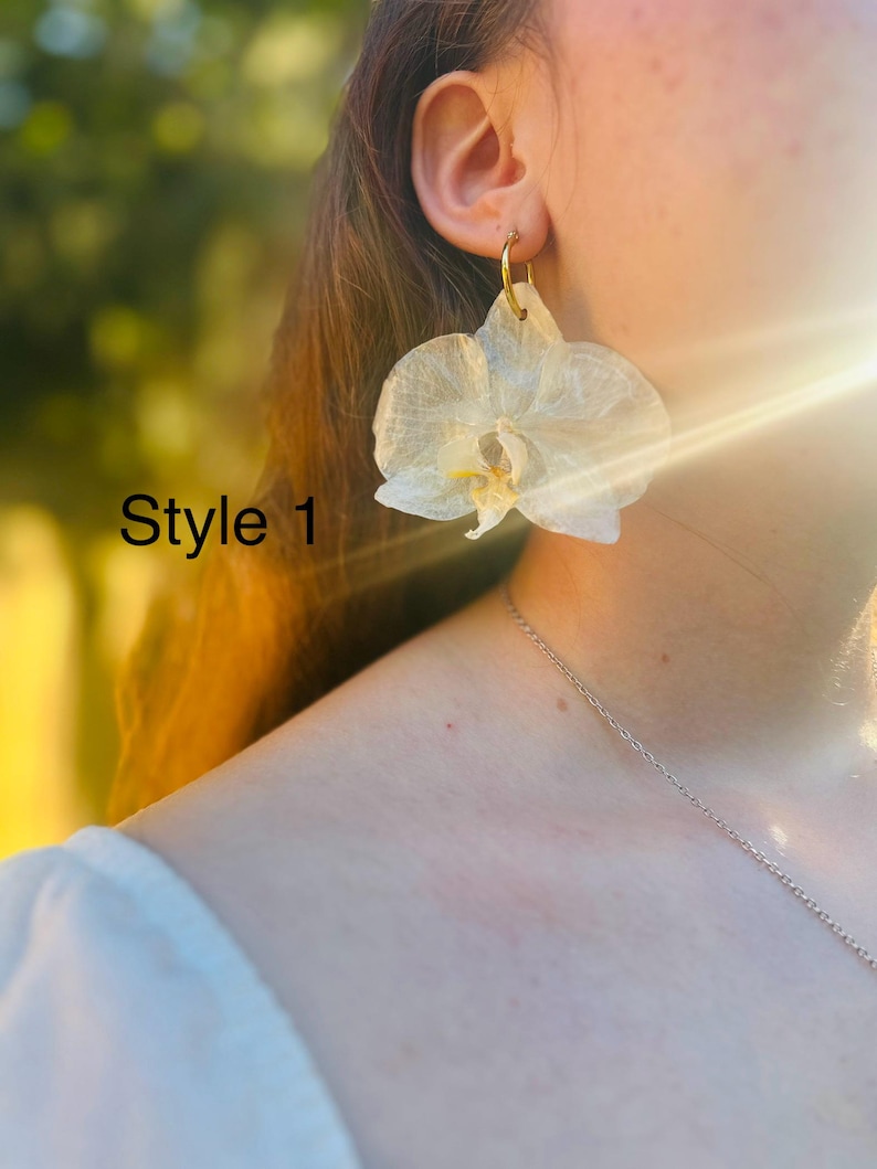 Pode incluir: Um brinco de flor de orqu&iacute;dea branca transl&uacute;cida com um gancho de argola de metal dourado. A estrutura das p&eacute;talas &eacute; vis&iacute;vel com nervuras delicadas e um centro amarelo. O texto Style 1 est&aacute; impresso em preto no lado esquerdo.