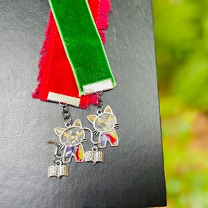 May include: Two velvet bookmarks, one red and one green, with silver cat charms attached. The charms feature a cat holding a book and have a floral design.