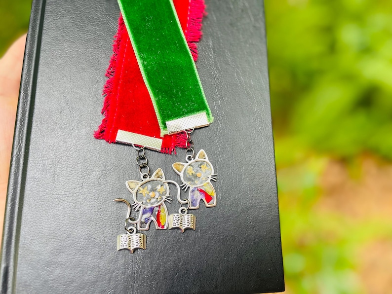 May include: Two velvet bookmarks, one red and one green, with silver cat charms attached. The charms feature a cat holding a book and have a floral design.