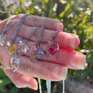 May include: Five silver necklaces with heart-shaped pendants featuring pressed flowers. The pendants are clear and show the flowers in detail. The necklaces are layered on a hand.