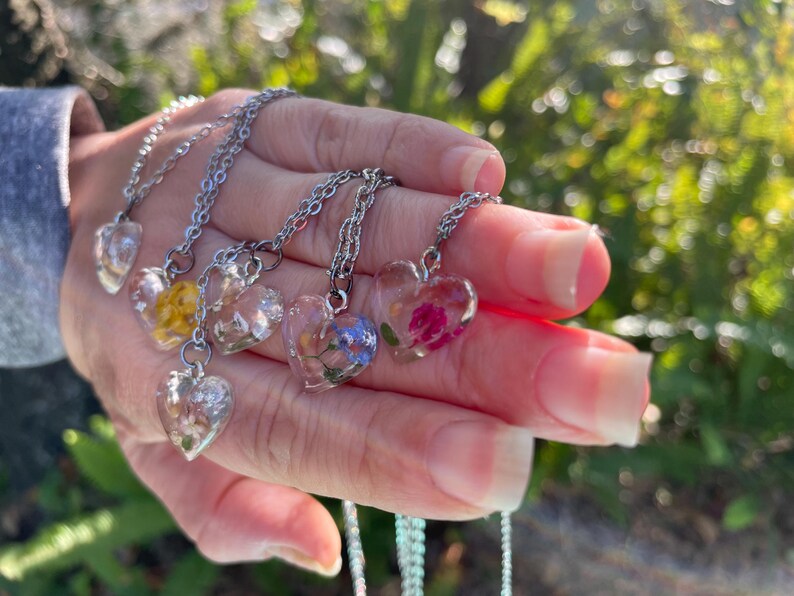 May include: Five silver necklaces with heart-shaped pendants featuring pressed flowers. The pendants are clear and show the flowers in detail. The necklaces are layered on a hand.