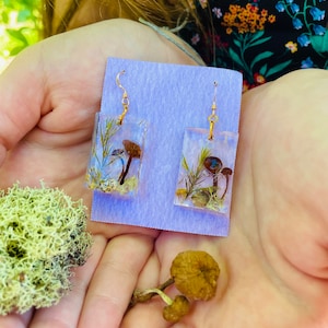 May include: A pair of gold earrings with rectangular resin charms featuring pressed mushrooms and greenery. The charms are on a purple background.