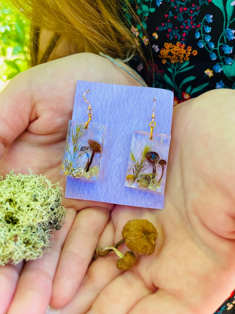 May include: A pair of gold earrings with rectangular resin charms featuring pressed mushrooms and greenery. The charms are on a purple background.