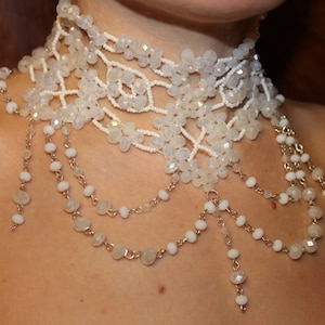 May include: A white beaded choker necklace with multiple strands of beads and a delicate chain design. The necklace features a layered look with a variety of bead sizes and shapes.