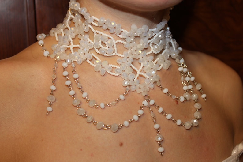 May include: A white beaded choker necklace with multiple strands of beads and a delicate chain design. The necklace features a layered look with a variety of bead sizes and shapes.