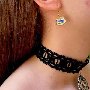 Puede incluir: Un pendiente dorado con forma de gato con fondo blanco y dise&ntilde;o floral azul. Tambi&eacute;n se ve un collar de gargantilla de encaje negro con un patr&oacute;n circular. La imagen muestra joyas.