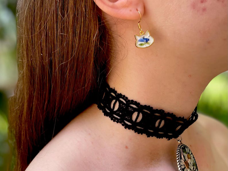Puede incluir: Un pendiente dorado con forma de gato con fondo blanco y dise&ntilde;o floral azul. Tambi&eacute;n se ve un collar de gargantilla de encaje negro con un patr&oacute;n circular. La imagen muestra joyas.