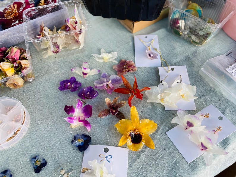 Pode incluir: Uma cole&ccedil;&atilde;o de pe&ccedil;as de joalharia feitas com flores de orqu&iacute;dea revestidas a resina, incluindo pendentes, brincos em cart&otilde;es brancos e acess&oacute;rios de cabelo. O sortido apresenta orqu&iacute;deas amarelas, roxas, vermelhas e brancas.