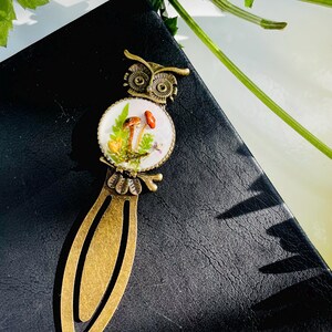 May include: A bronze owl bookmark with a round glass charm featuring a white background with a miniature mushroom scene. The charm is surrounded by a bronze frame.