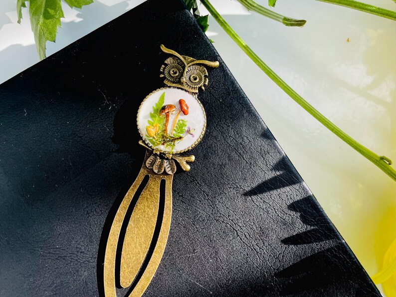 May include: A bronze owl bookmark with a round glass charm featuring a white background with a miniature mushroom scene. The charm is surrounded by a bronze frame.
