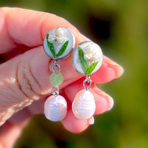 May include: A pair of silver dangle earrings with a white pearl charm and a white and green flower charm. The flower charm is made of resin and features a pressed flower.