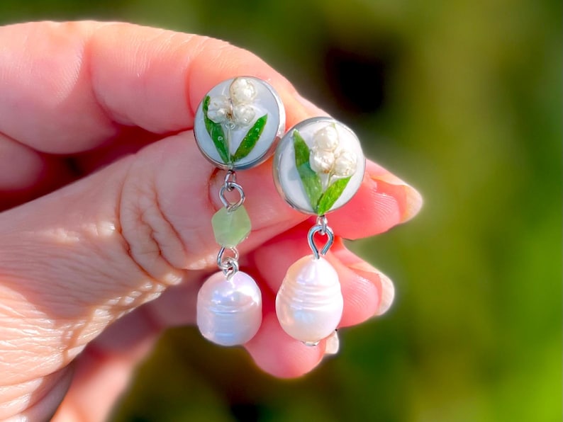 May include: A pair of silver dangle earrings with a white pearl charm and a white and green flower charm. The flower charm is made of resin and features a pressed flower.