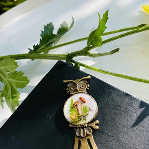 May include: A bronze owl bookmark with a white porcelain owl design featuring mushrooms and green foliage. The owl's eyes are black and the bookmark has a long, thin metal tail.