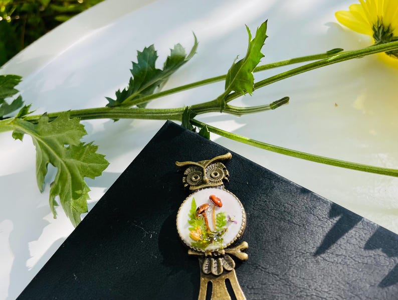 May include: A bronze owl bookmark with a white porcelain owl design featuring mushrooms and green foliage. The owl's eyes are black and the bookmark has a long, thin metal tail.