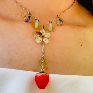 May include: A silver chain necklace with a delicate flower charm and a red strawberry charm. The flower charm is made of resin and features a white flower with green leaves. The strawberry charm is made of resin and is a realistic representation of a strawberry.