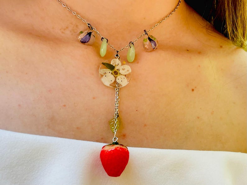 May include: A silver chain necklace with a delicate flower charm and a red strawberry charm. The flower charm is made of resin and features a white flower with green leaves. The strawberry charm is made of resin and is a realistic representation of a strawberry.
