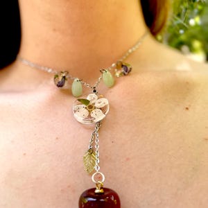 May include: A silver chain necklace with a red glass apple pendant. The apple is attached to a smaller pendant with a white flower inside a clear resin circle. The necklace also has green and purple glass beads.