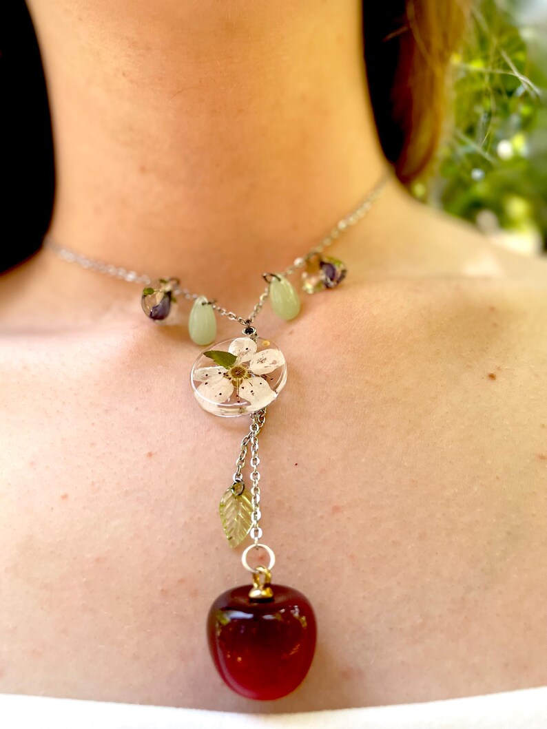 May include: A silver chain necklace with a red glass apple pendant. The apple is attached to a smaller pendant with a white flower inside a clear resin circle. The necklace also has green and purple glass beads.