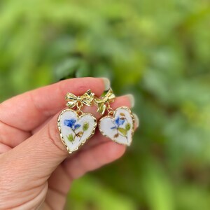 K&ouml;nnte beinhalten: Ein Paar goldene herzf&ouml;rmige Ohrringe mit wei&szlig;em Hintergrund und gepressten blauen und gr&uuml;nen Blumen darin. Die Ohrringe haben einen goldenen Schleifenanh&auml;nger.