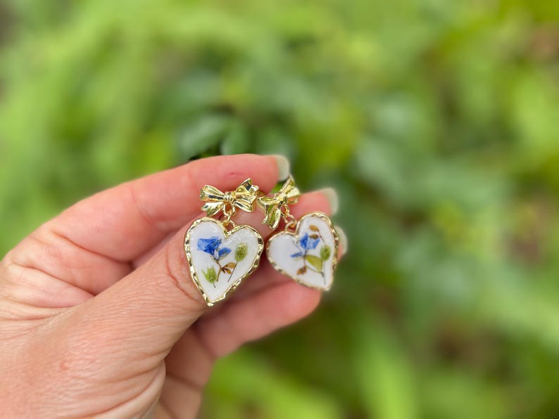 K&ouml;nnte beinhalten: Ein Paar goldene herzf&ouml;rmige Ohrringe mit wei&szlig;em Hintergrund und gepressten blauen und gr&uuml;nen Blumen darin. Die Ohrringe haben einen goldenen Schleifenanh&auml;nger.