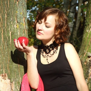 May include: A woman with brown hair and a black dress holds a red apple in her hand. She is wearing a black beaded necklace and a red scarf. The woman is standing in front of a tree trunk.
