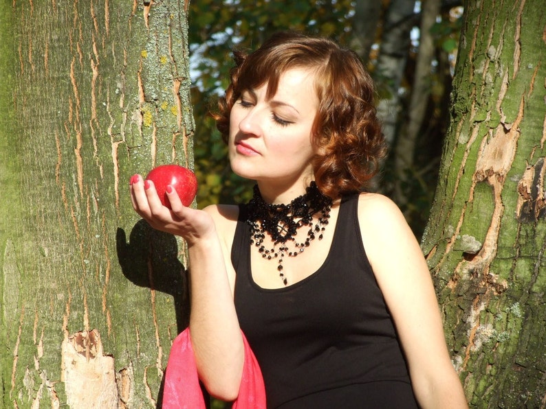 May include: A woman with brown hair and a black dress holds a red apple in her hand. She is wearing a black beaded necklace and a red scarf. The woman is standing in front of a tree trunk.