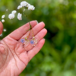 Puede incluir: Un par de pendientes de aro dorados con un dise&ntilde;o de resina transparente con flores moradas y blancas prensadas.