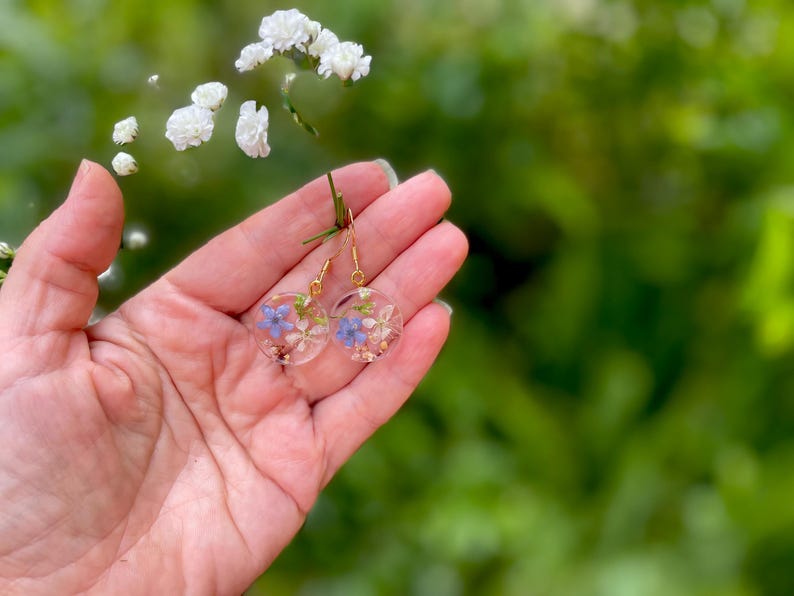 Puede incluir: Un par de pendientes de aro dorados con un dise&ntilde;o de resina transparente con flores moradas y blancas prensadas.