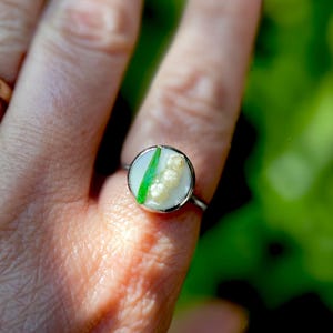 May include: A silver ring with a white and green resin inlay featuring a cluster of white flowers. The ring is on a person's finger.