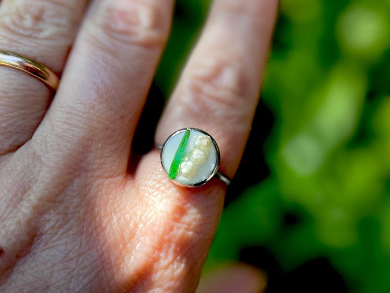 May include: A silver ring with a white and green resin inlay featuring a cluster of white flowers. The ring is on a person's finger.