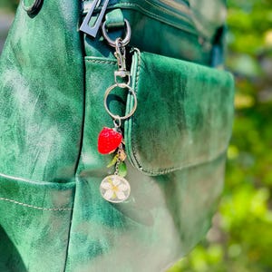Peut inclure: Un sac &agrave; dos en cuir vert avec une fermeture &eacute;clair argent&eacute;e et un porte-cl&eacute;s d&eacute;coratif. Le porte-cl&eacute;s comprend un charm en forme de fraise rouge, un petit charm dor&eacute;, une perle verte et un charm circulaire transparent avec une fleur blanche.