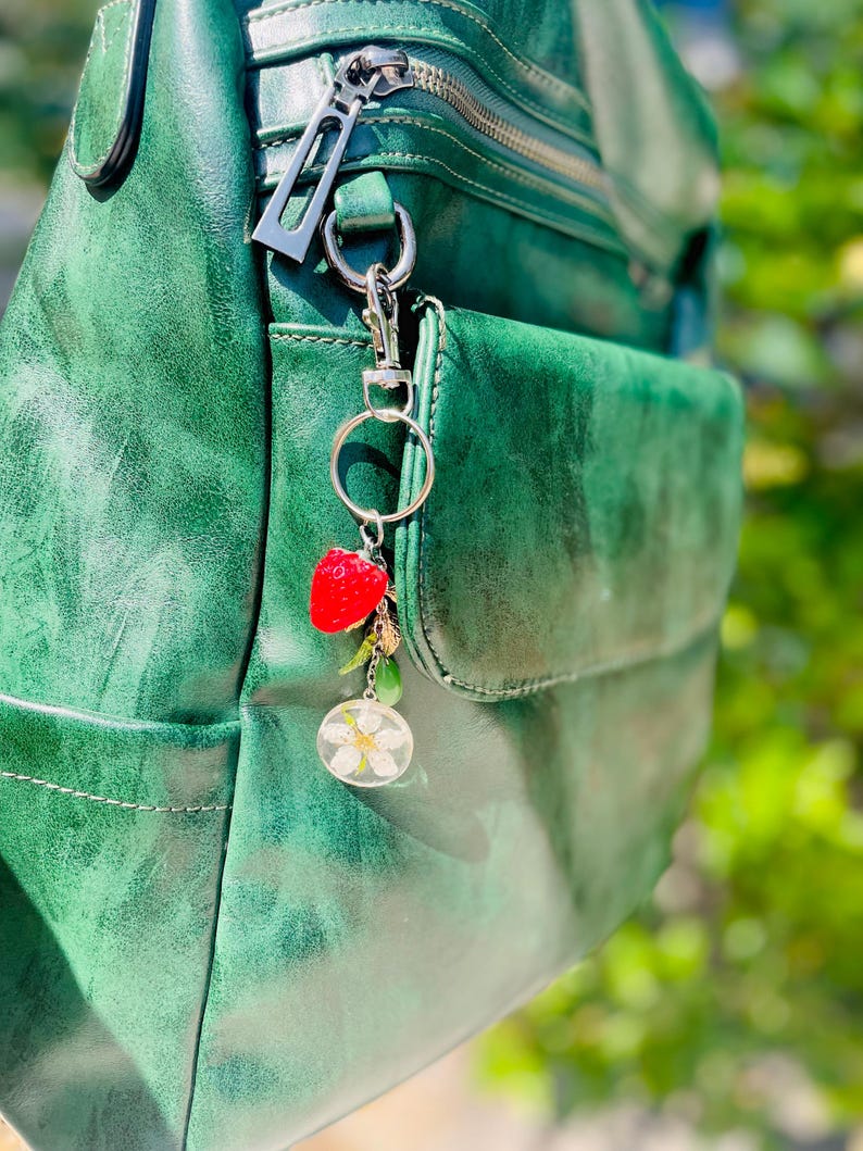 Peut inclure: Un sac &agrave; dos en cuir vert avec une fermeture &eacute;clair argent&eacute;e et un porte-cl&eacute;s d&eacute;coratif. Le porte-cl&eacute;s comprend un charm en forme de fraise rouge, un petit charm dor&eacute;, une perle verte et un charm circulaire transparent avec une fleur blanche.