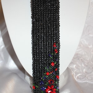 May include: A black beaded necklace with a tie-like design. The necklace features a cascade of black beads, with a floral pattern of red, green, and blue beads at the bottom. The necklace is displayed on a white mannequin.