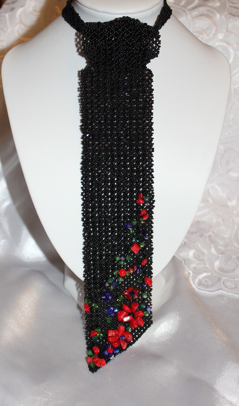 May include: A black beaded necklace with a tie-like design. The necklace features a cascade of black beads, with a floral pattern of red, green, and blue beads at the bottom. The necklace is displayed on a white mannequin.