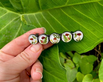 May include: A black metal barrette featuring five clear, circular resin domes. Each dome contains a miniature mushroom and plant arrangement. The mushrooms have red caps and brown stems, with green ferns and other plants. The barrette is held against a large green leaf.