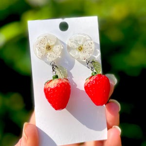 Op de afbeelding: Een paar oorbelletjes met een witte bloemharsbedel en een rode aardbeienbedel.