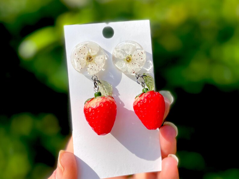 Op de afbeelding: Een paar oorbelletjes met een witte bloemharsbedel en een rode aardbeienbedel.