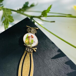 May include: A bronze bookmark featuring an owl design. The owl is holding a mushroom in its beak. The bookmark has a white enamel background with a mushroom illustration.