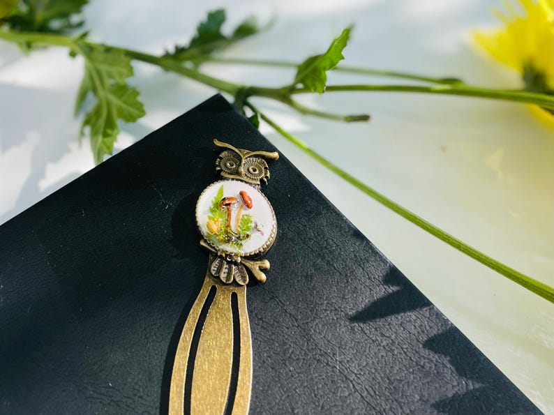 May include: A bronze bookmark featuring an owl design. The owl is holding a mushroom in its beak. The bookmark has a white enamel background with a mushroom illustration.