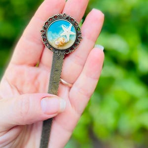 May include: A bronze metal bookmark with a decorative circular top featuring a miniature beach scene. The scene includes a white starfish, seashells, and blue water. The bookmark has a ruler design along its length.