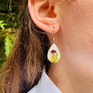 Puede incluir: Un par de pendientes de gota de oro con fondo blanco y un peque&ntilde;o hongo rojo con vegetaci&oacute;n verde en su interior.