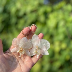 Pode incluir: Um par de brincos com p&eacute;talas de orqu&iacute;dea transl&uacute;cidas em tom branco-sujo, presas a argolas circulares douradas. As p&eacute;talas delicadas e de formato org&acirc;nico t&ecirc;m um brilho subtil e uma apar&ecirc;ncia suave e em camadas, suspensas por an&eacute;is de metal.