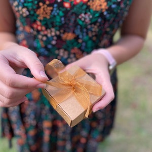 Puede incluir: Una peque&ntilde;a caja de regalo marr&oacute;n con una cinta dorada atada en un lazo. La caja est&aacute; siendo abierta por una mano.