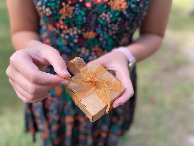 Puede incluir: Una peque&ntilde;a caja de regalo marr&oacute;n con una cinta dorada atada en un lazo. La caja est&aacute; siendo abierta por una mano.