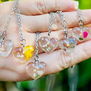 Puede incluir: Cinco collares de plata con colgantes en forma de coraz&oacute;n rellenos de flores prensadas. Cada colgante presenta una disposici&oacute;n diferente de flores, incluidas flores blancas, amarillas, rosas y azules.